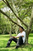 Young man sitting under tree, listening to music - Asia Images Group