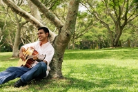 Young man sitting under tree, playing guitar - Asia Images Group