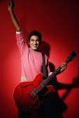 Young man holding red guitar, standing against red wall, smiling at camera - Asia Images Group