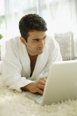 Man in bathrobe, lying on floor at home, using laptop - Asia Images Group
