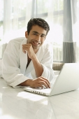 Man in bathrobe, using laptop, hand on chin, smiling at camera - Asia Images Group