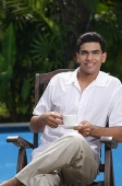 Man sitting in chair by the pool, holding a cup and looking at camera - Asia Images Group
