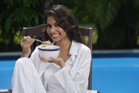 Woman in chair by the pool, holding bowl of cereal and looking at camera - Asia Images Group