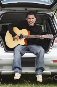 Young man sitting car boot playing guitar - Asia Images Group