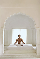 man meditating on bed in white room - Alex Mares-Manton