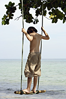 Young boy standing on swing by ocean - Alex Mares-Manton