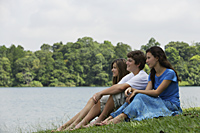 Three teens sitting on grass next to lake - Nugene Chiang