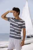 Young man looking out to ocean, standing in front of sail boat - Alex Mares-Manton