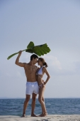 man holding giant leaf over woman as they stand on beach - Alex Mares-Manton
