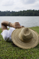 Young man lying on grass near lake, hat covering face - Nugene Chiang